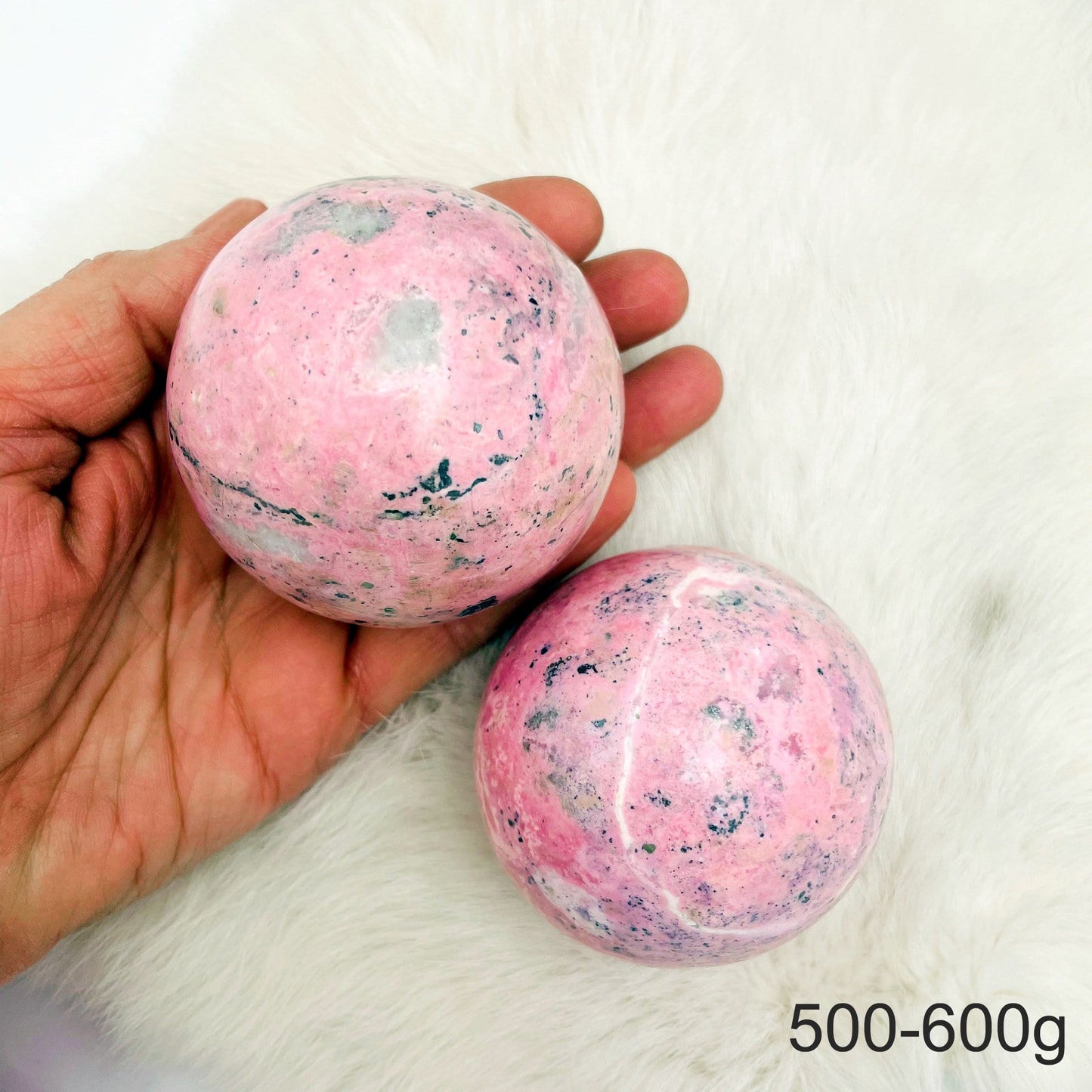 Hand holding 1 500-600g Rhodonite Polished Sphere with another one on fur backgorund