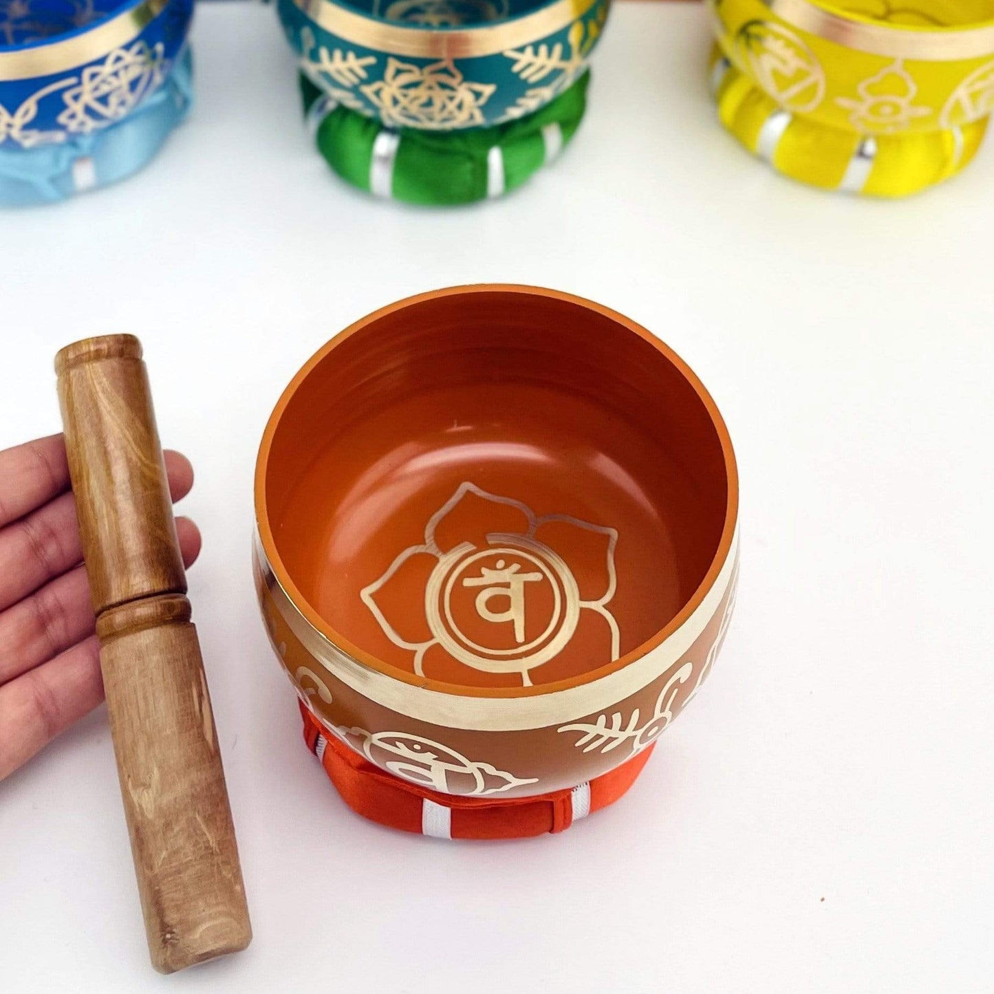 Orange Brass Tibetan Chakra Singing Bowl on a pillow next to a hand holding a mallot