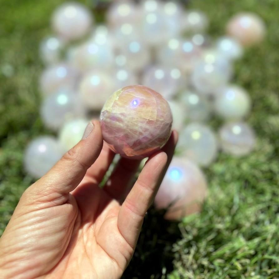 Hand holding up Rose Quartz Angel Aura Titanium Spheres with others blurred on grassy backround