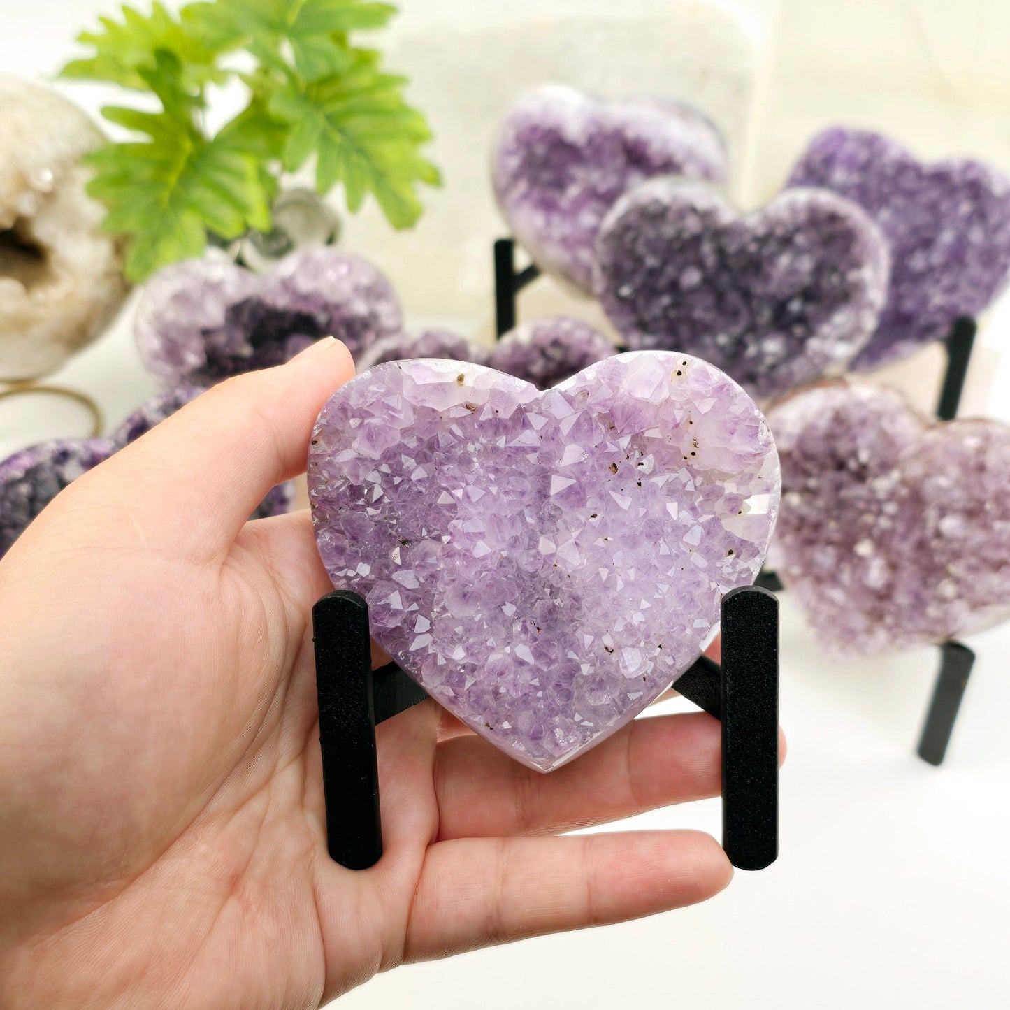 Amethyst Crystal Heart on Metal Tripod Stand - in hand for size reference with other amethyst hearts on metal stands in the background with props and plants