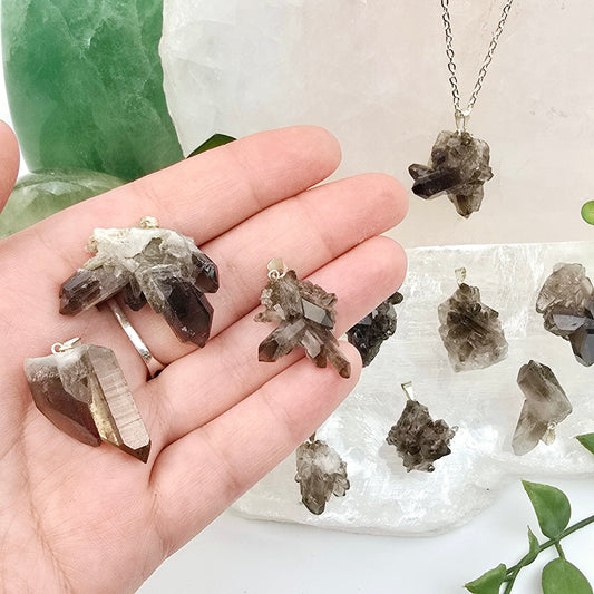 Smokey Quartz Rough Crystal Cluster Pendant with Silver Bail - three in hand for size reference with others in the background one on silver chain with props and plants