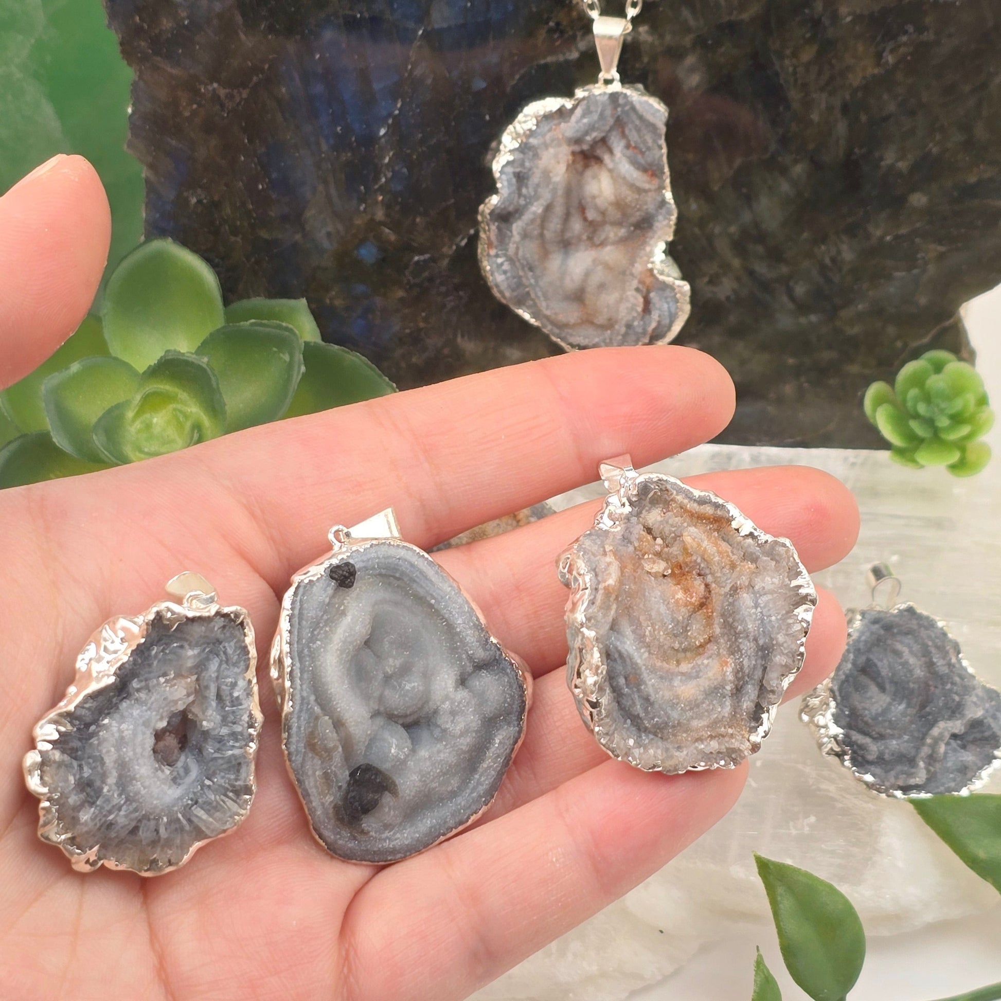 Chalcedony Galaxy Druzy Pendant with Silver Plated Edge and Bail - three in hand for size reference with others in the background on chain and selenite platter with props and plants