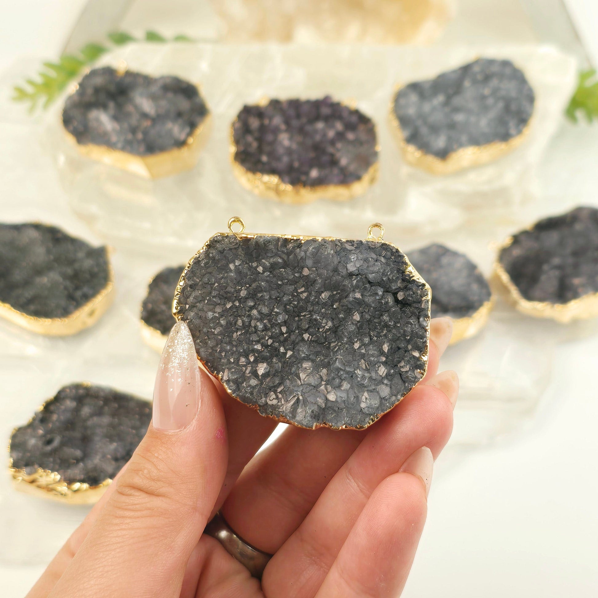 Gray Amethyst Druzy Oval Slab Pendant with 24k Gold Electroplated Edge and Double Bail - one in hand for size reference with others in the background on selenite platters