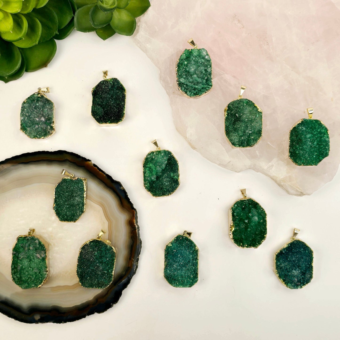 Green Druzy Freeform Pendant with 24k Gold Electroplated Edge and Bail twelve on crystal platters and white surface with plants in the background