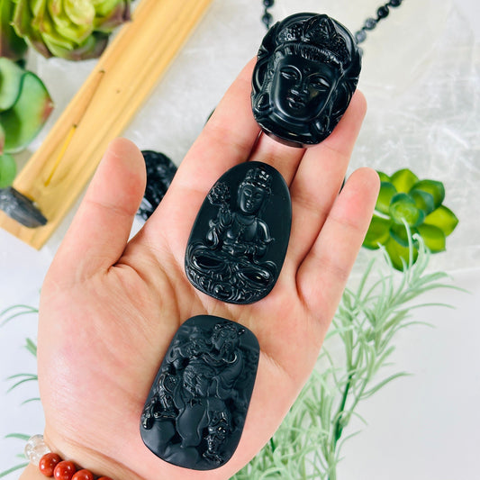Black Obsidian Buddha Head, Sitting Buddha, or Guan Yu Carved Crystal Pendant - You Choose Style - all variants in hand with props and plants in the background
