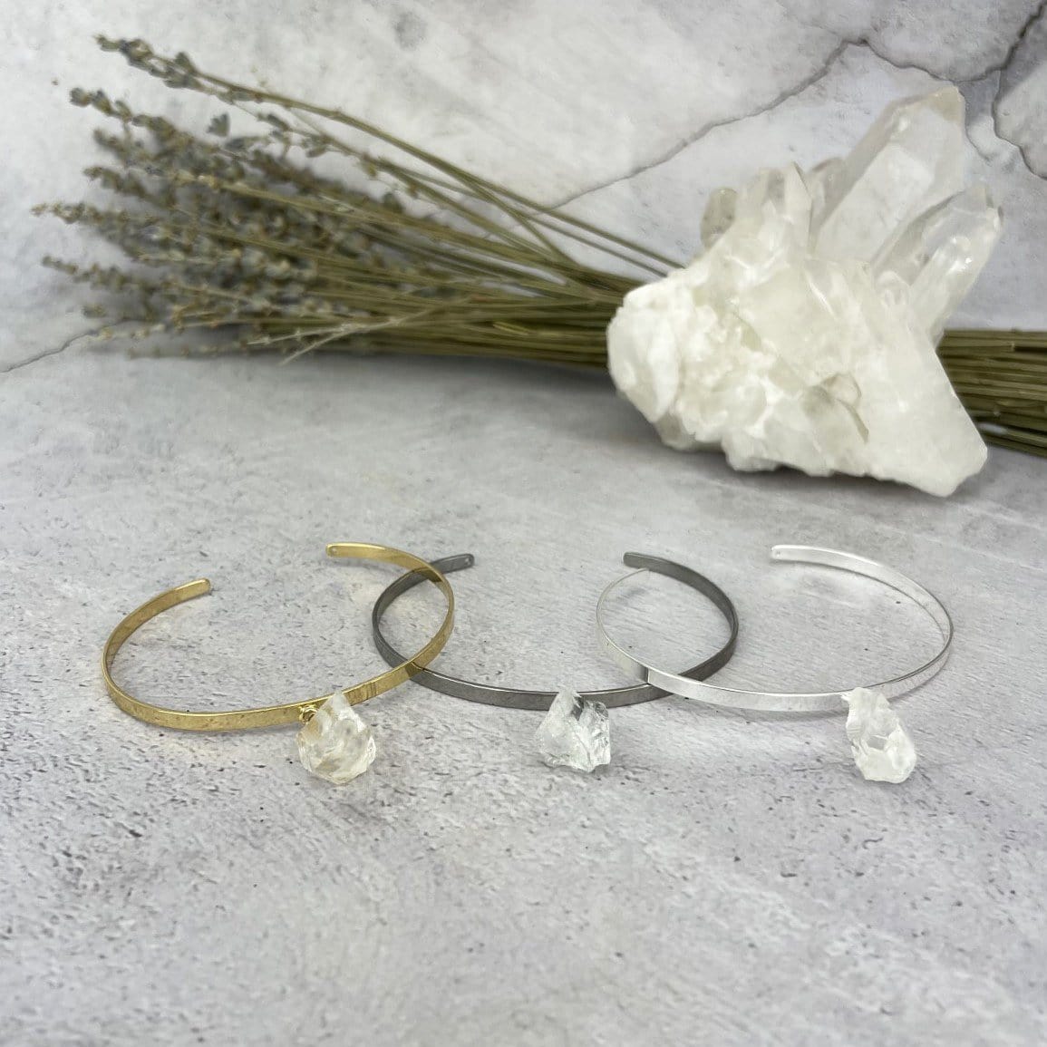 Gold and silver bracelets that are thin metal with a rough crystal quartz hanging from the middle.  Pictured here on a purple and white point. Pictured here in gold plated, gun metal plated, and silver plated on a cement background.