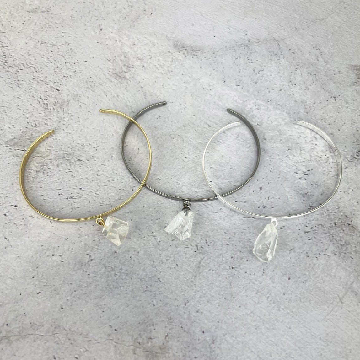 Gold and silver bracelets that are thin metal with a rough crystal quartz hanging from the middle.  Pictured here on a purple and white point. Pictured here in gold plated, gun metal plated, and silver plated on a cement background.