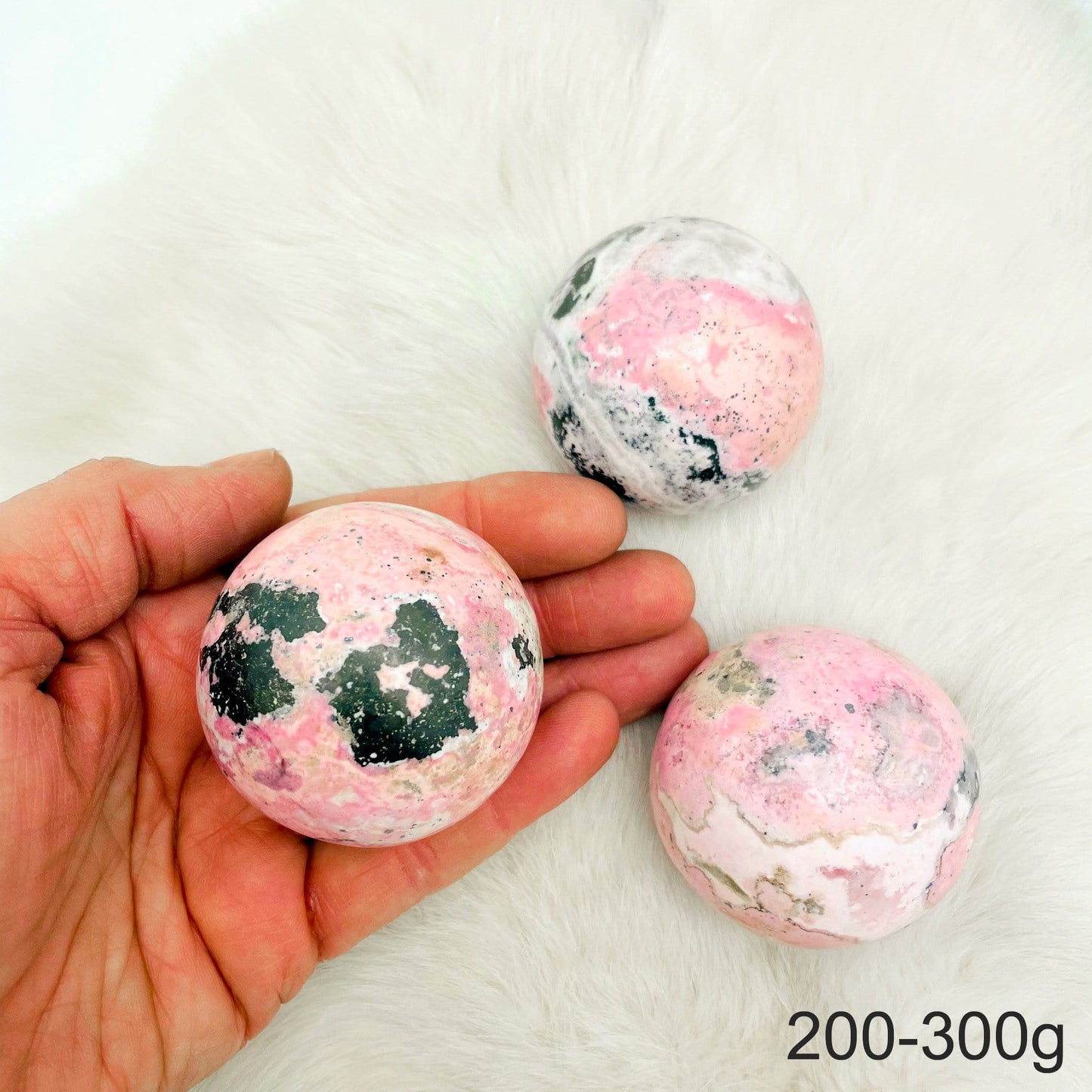 Hand holding up 1 200-300g Rhodonite Polished Sphere with 2 others on fur background