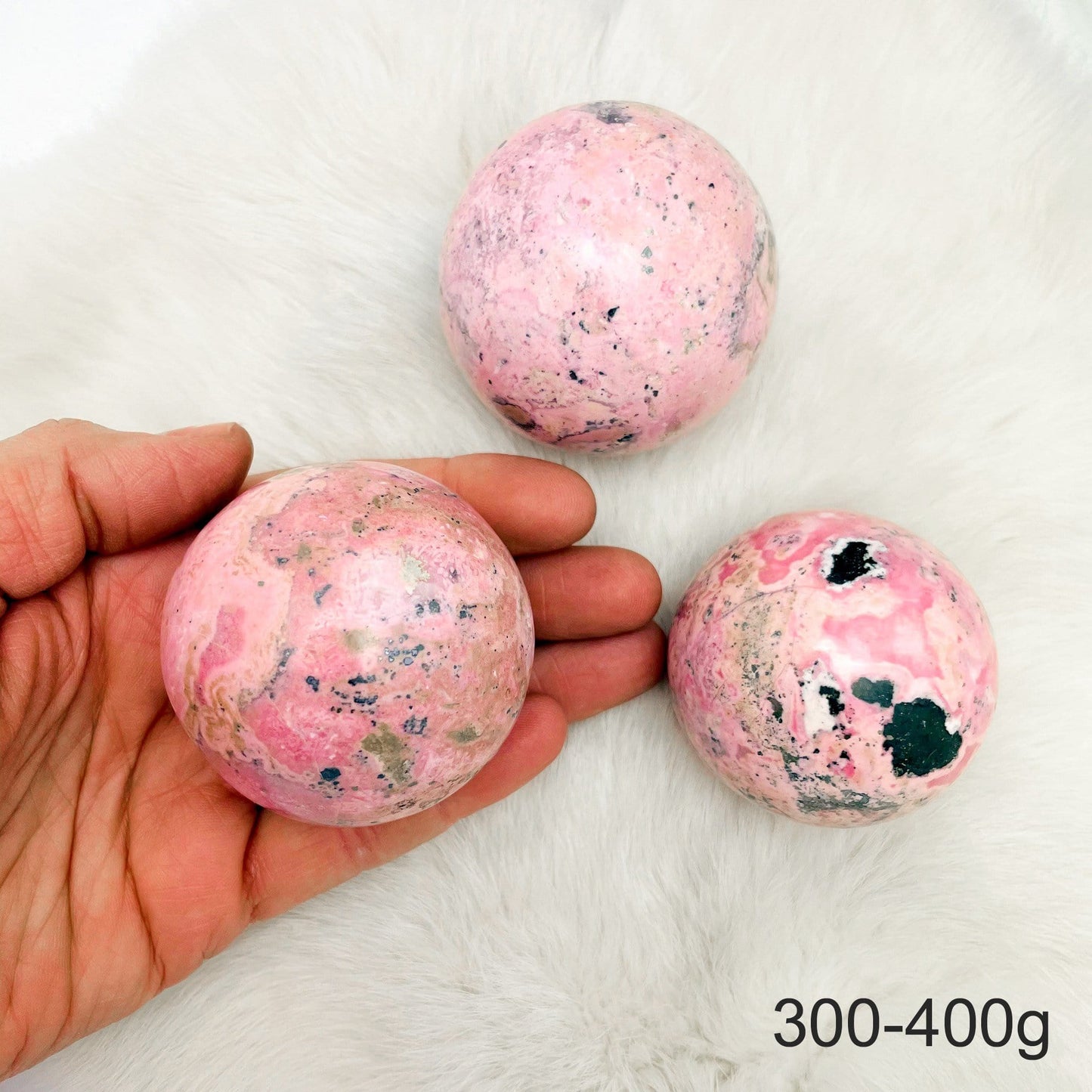 Hand holding up 1 300-400g Rhodonite Polished Sphere with 2 others on fur background