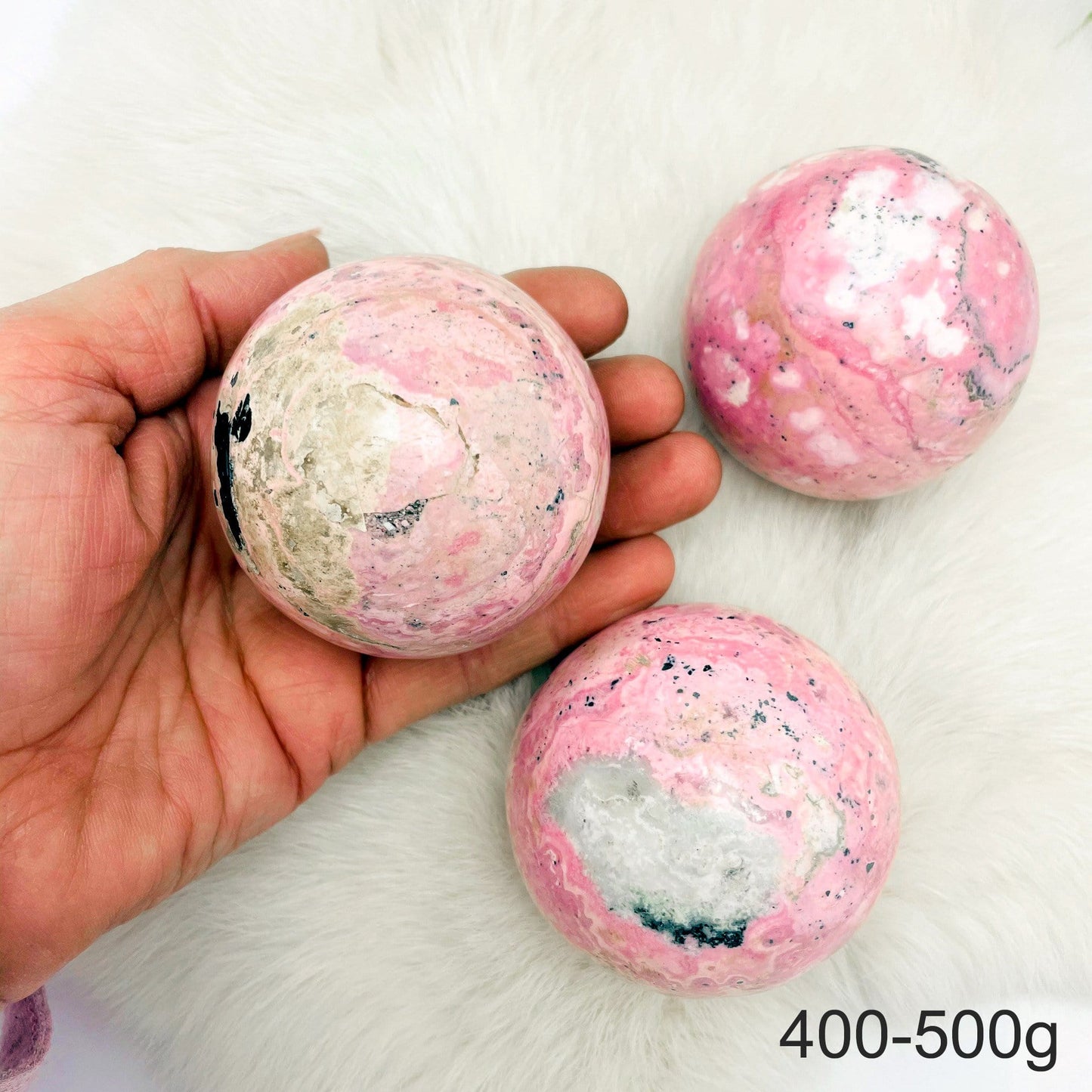 Hand holding up 1 400-500g Rhodonite Polished Sphere with 2 others on fur background
