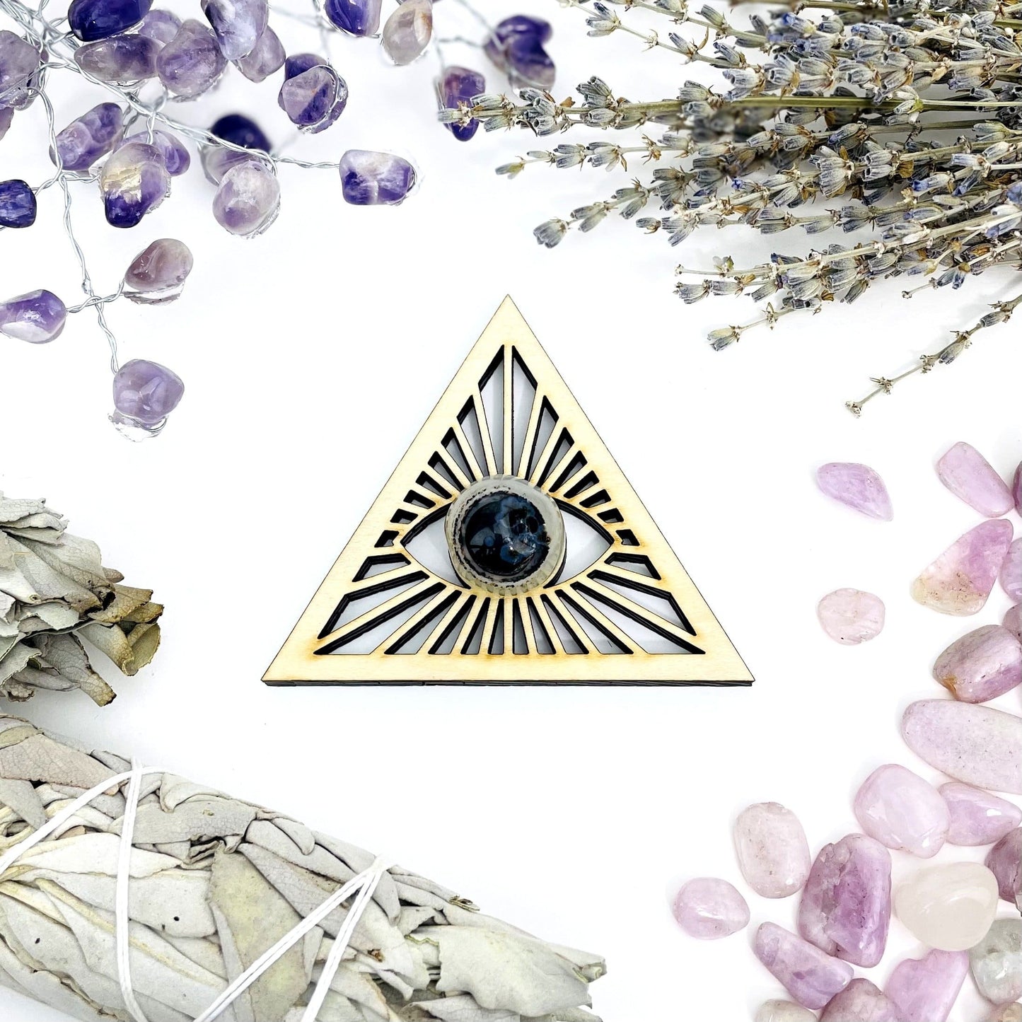 A Eye of Providence Wooden Sphere Stand in middle of tumble stones and a bundle of sage.