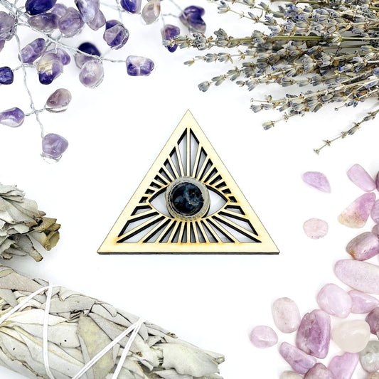 A Eye of Providence Wooden Sphere Stand in middle of tumble stones and a bundle of sage.