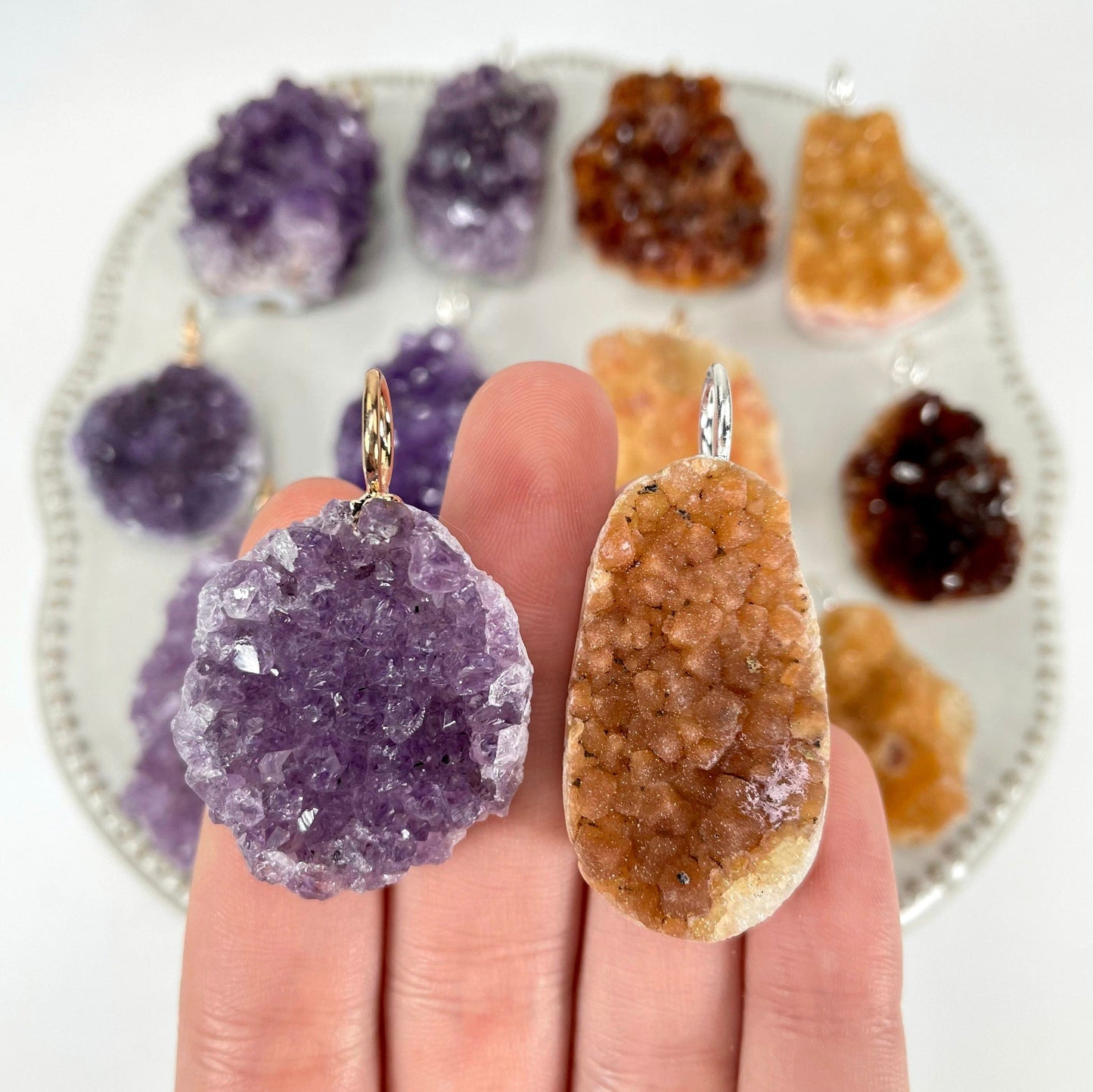 close up of two amethyst druzy cluster pendant options in hanf for size reference and possible variations with many others on display in the background