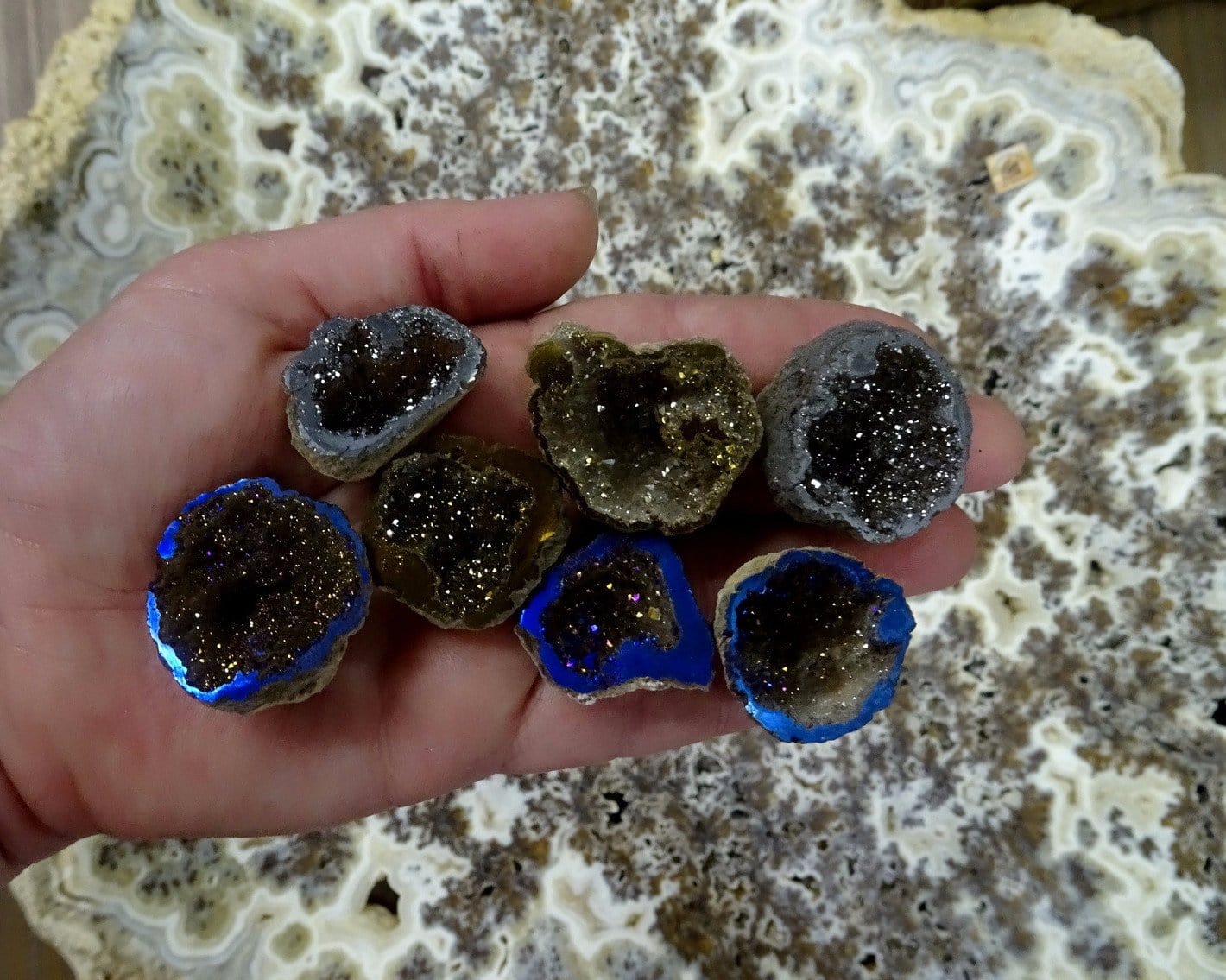 hand holding up 7 mystic half geodes in platinum, gold, and rainbow in front of a crystal